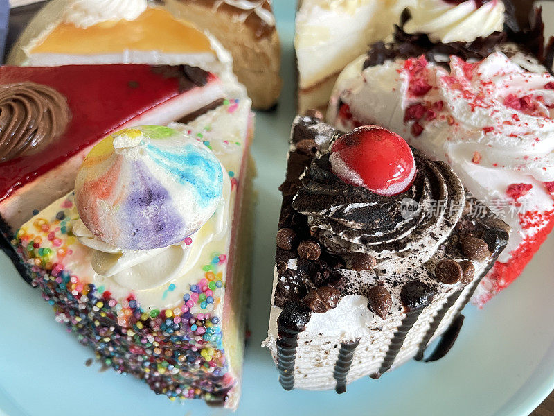 粉彩蓝色盘子上各种蛋糕切片的全帧图像，天使食物蛋糕，巧克力和樱桃蛋糕，黑森林蛋糕，红丝绒蛋糕，彩虹蛋糕，咖啡和大理石巧克力蛋糕，咸味焦糖蛋糕，专注于前景，抬高视图