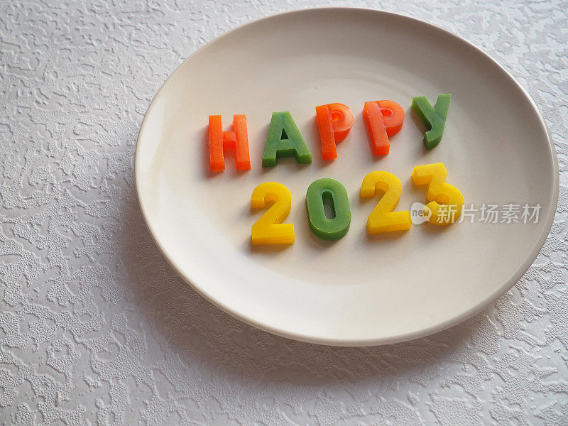 快乐的2023年。新年快乐。新年的明信片。白色的盘子上排列着橙色、绿色、黄色的字母和数字。平的。水平面上的物体