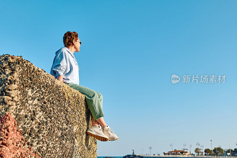 年轻女子坐在海边的大石头上，背景是蓝天，享受自由。女旅行者在宁静的大自然中放松。心理健康，幸福，旅行冒险和健康的生活方式。