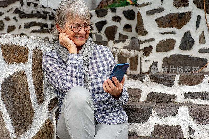 快乐的老年妇女坐在室外楼梯上，从智能手机上得到一些好消息。快乐的老妇人使用手机。一位漂亮的老妇人收到了家人的电话留言