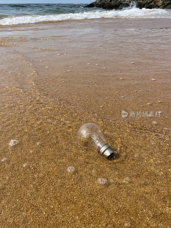 近距离图像玻璃灯泡被冲到沙滩上在退潮水的边缘，破碎的海浪冲刷海洋垃圾和岸上的污染，乱扔的沙子，肮脏的海滩，关注前景