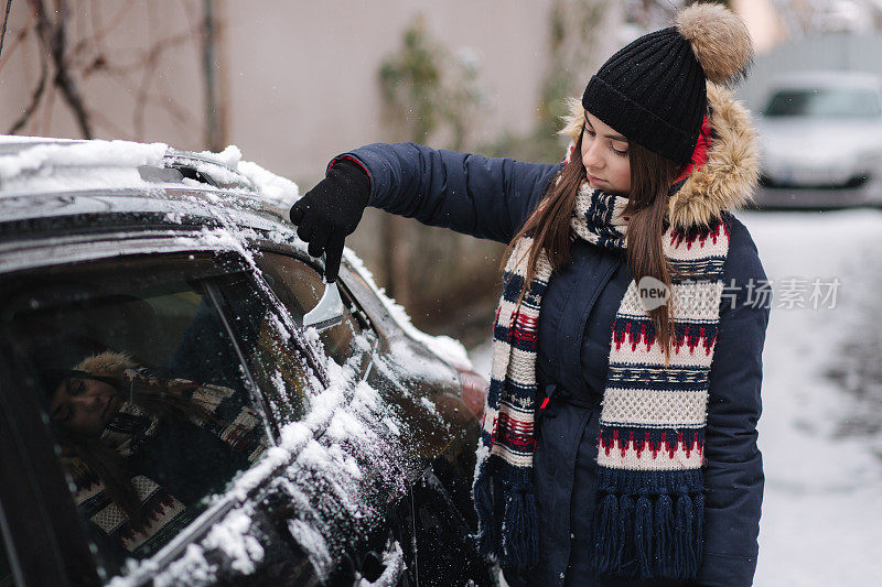 一位妇女正在用刮雪机清理汽车上被雪覆盖的窗户。漂亮的女人穿着暖和，在户外打扫她的车。寒冷的雪和霜的早晨。黑色的车