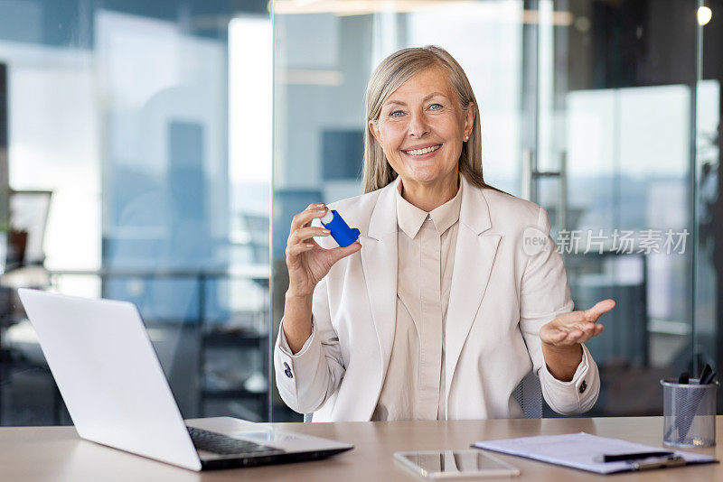 在现代办公室里，成熟的白人女人对着相机微笑，拿着吸入器，用台式机和笔记本电脑。阳性女士在工作时间服用医生处方的药物预防哮喘发作