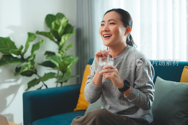 亚洲妇女坐在客厅的沙发上用吸管喝凉水休息。