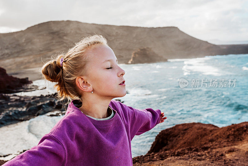 年轻的女孩闭着眼睛享受着吹在脸上的海风。微笑的金发女孩享受加那利群岛的大自然。和孩子们一起去海滩度假的概念。
