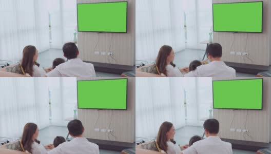 亚洲幸福家庭坐在家里的客厅看电视，一起度过宝贵的时间，频道到绿色屏幕显示器为您的信息高清在线视频素材下载