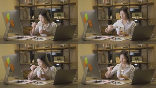 亚洲自由职业者精明的商业女性一边吃着方便面，一边在家里的客厅里用笔记本电脑工作。高清在线视频素材下载