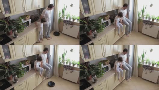 父亲和儿子抬起他们的腿，让机器人真空吸尘器清理厨房的地板高清在线视频素材下载