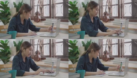 快乐的年轻亚洲妇女微笑与手机，同时使用笔记本电脑在家里工作高清在线视频素材下载