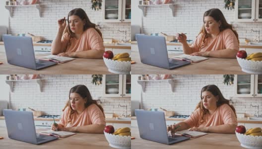 年轻的胖女人在笔记本电脑上看网络研讨会，做笔记，坐在家里的厨房里高清在线视频素材下载