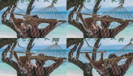 在蓝色海洋前的海滩上，一个男人在吊床上放松地摇摆着。把他藏在巫师树的阴影下躲避阳光。高清在线视频素材下载