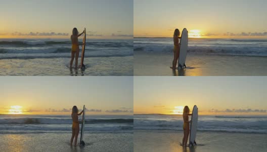 美丽的年轻女子在泳衣站在海里，同时持有冲浪板。大海浪和日落的背景。高清在线视频素材下载