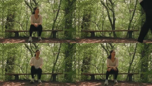 体育训练。一位身穿运动服的年轻女子坐在森林公园的长椅上慢跑，戴着耳机听着音乐。实时高清在线视频素材下载