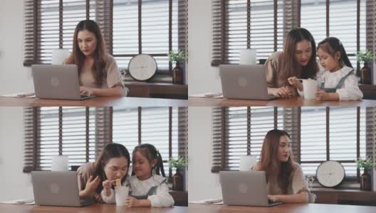 自由职业者亚洲母亲在桌子上工作与她的笔记本电脑，她的女儿喂她的面条在客厅在家里，幸福的家庭人的生活理念。高清在线视频素材下载