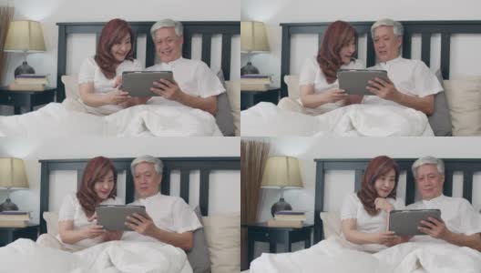 亚洲老年夫妇在家里使用平板电脑。亚洲老爷爷老爷爷，夫妻高兴地醒来后，躺在家里卧室的床上看电影的概念。缓慢的运动。高清在线视频素材下载