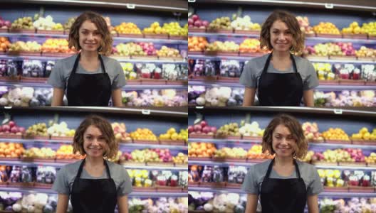 年轻漂亮的女售货员穿着黑色围裙站在超市货架上的水果在背景，看着相机和微笑。商贸以人为本的理念高清在线视频素材下载