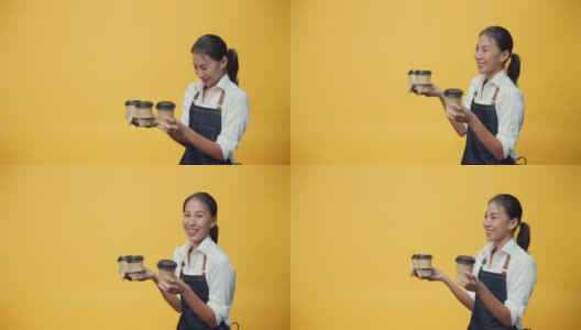 快乐微笑的亚洲女士企业老板穿着围裙友好地看广告文案空间持有咖啡或茶孤立在黄色的背景。高清在线视频素材下载