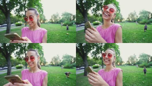 微笑的女人与滑稽的脸和粉红色的眼镜站在日落城市绿色公园的背景和使用手机高清在线视频素材下载