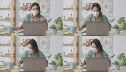 亚洲女商人戴着医用口罩，使用笔记本电脑与同事在视频通话中讨论计划，同时在家起居室工作。高清在线视频素材下载
