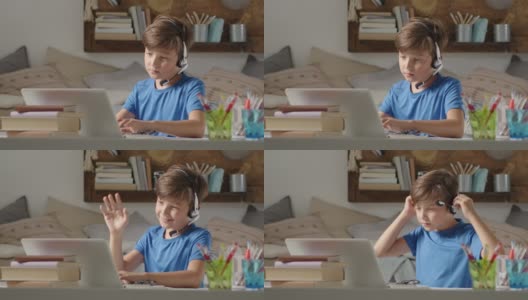 微笑的学生男孩在家学习，远程学校连接到网络上的电脑，举起他的手回答问题或问候他的同学，坐在摆满书的书桌前，戴着耳机高清在线视频素材下载