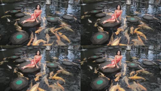迷人的深色皮肤的模特穿着红色的衣服，放松在石径上的池塘五颜六色的鱼高清在线视频素材下载