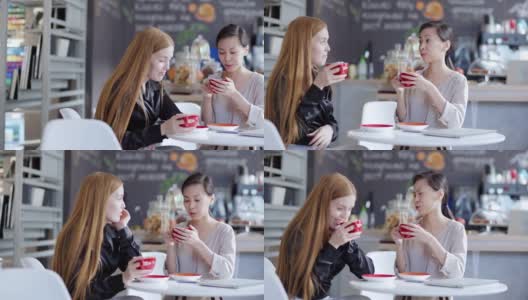 右图是两个年轻女人坐在咖啡馆的桌子旁，喝着咖啡聊天。亚洲女性讲述故事情绪化的红发白人朋友高清在线视频素材下载