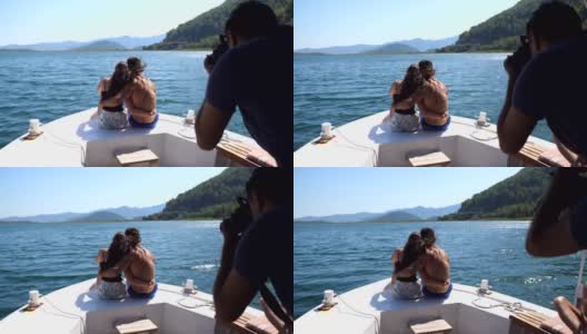 一对不认识的年轻夫妇坐在船头，对着不认识的摄影师摆姿势。幸福的情侣在船上共度时光，享受夏天的旅行。假期的概念。后方后视图高清在线视频素材下载