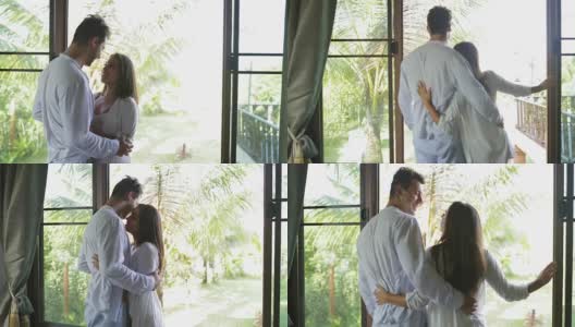 年轻夫妇看着阳台拥抱，美丽的女孩和男人在卧室早上高清在线视频素材下载