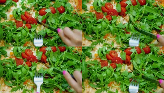 特写，大而多汁的热披萨，配上绿色蔬菜，芝麻菜和樱桃番茄。女性用刀叉切披萨高清在线视频素材下载
