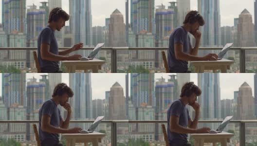 一个年轻的自由职业者在阳台上用笔记本电脑工作，背景是一个摩天大楼林立的城市中心。自由职业者it专家解决客户的问题高清在线视频素材下载