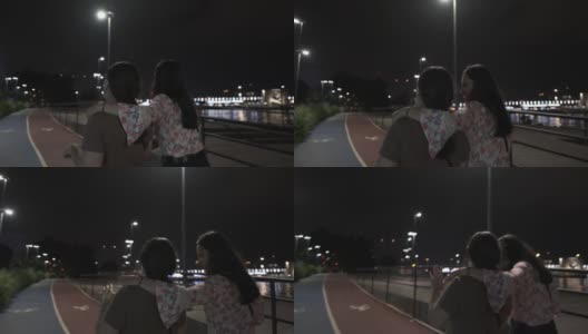 跟踪跟踪摄像头拍摄的两个快乐的姐妹笑着微笑着走在海滨地区的步行街在灯泡下在夏天的夜晚，一位女士把她的手臂放在另一个女人的肩膀4K分辨率高清在线视频素材下载