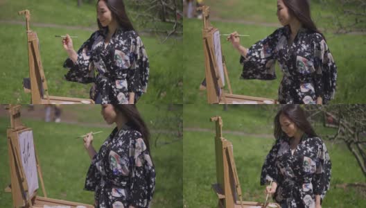 旁边的亚洲女人穿着和服，用毛笔在画布上微笑。这是一幅灵感迸发、技艺精湛的日本画家在户外享受阳光明媚的春日的肖像。缓慢的运动。高清在线视频素材下载