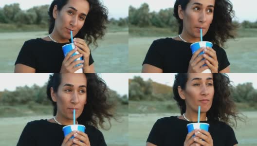 一个可爱的黑发女孩抿着蓝色玻璃杯里的饮料，看着梦中的日落。高清在线视频素材下载