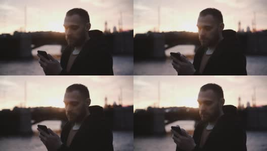 年轻的大胡子男子花时间在美丽的河堤上的肖像，在傍晚日落时使用智能手机高清在线视频素材下载
