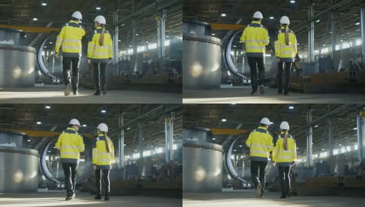 男工程师和女工程师在重工业制造厂边走边讨论的背影照片。大型金属制品结构，管道元素躺在周围。高清在线视频素材下载