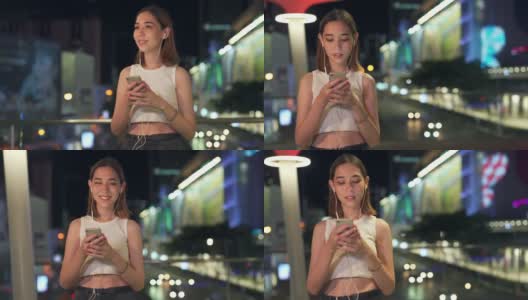 年轻亚洲女人的4K肖像走在城市的街道听音乐从耳机在晚上高清在线视频素材下载
