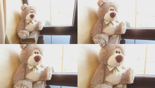 泰迪熊坐在窗台上，可爱的泰迪熊靠近窗户，在她的脖子上有一只蝴蝶，浅棕色的，儿童的软玩具，一个泰迪熊在她的脖子上有一只蝴蝶高清在线视频素材下载