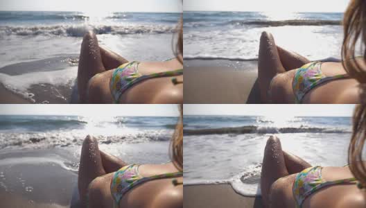 难以辨认的性感女孩躺在海滩的波浪中。美丽的年轻女子放松在海边的暑假旅行。海浪冲刷着晒黑的女性身体。慢动作特写后视图高清在线视频素材下载