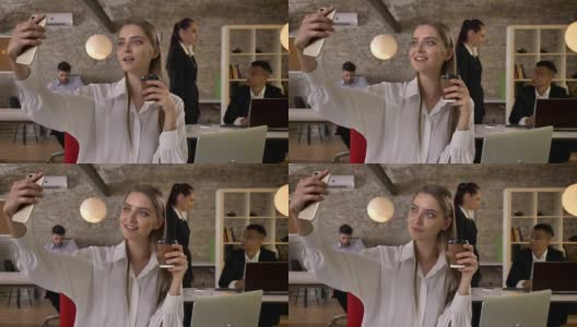 年轻迷人的女商人在办公室用智能手机端着咖啡自拍，她的同事们用科技、工作理念、商业理念、沟通理念进行网络交流高清在线视频素材下载