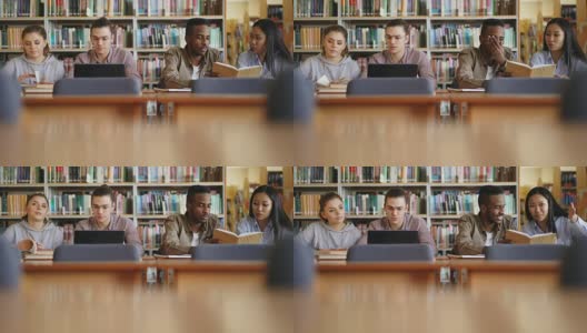 多民族学生坐在图书馆里，桌上放着书和笔记本电脑，一起笑着准备考试高清在线视频素材下载