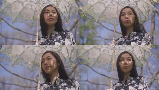 自信的年轻亚洲女性肖像，站在夏日公园，撑着太阳伞，环顾四周，享受大自然的美丽。美丽苗条的千禧一代在阳光明媚的春天花园里休息。高清在线视频素材下载