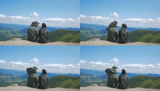 年轻貌美的女旅行者和年轻貌美的男坐在山顶上，眺望远处雄伟的森林。一对年轻相爱的游客坐在山脚下的岩石上拥抱在一起高清在线视频素材下载