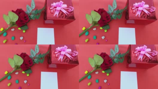 一个礼品盒系蝴蝶结丝带和红玫瑰和一张空白贺卡在红色背景的桌子上。平的。直接上图。设计情人节，周年纪念或节日庆祝装饰高清在线视频素材下载