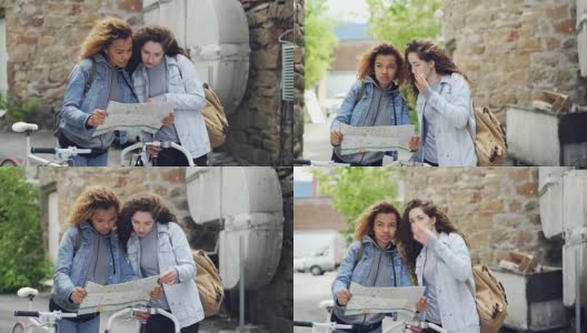 漂亮的女孩，非洲裔美国人和白人，看着地图站在未知的城市，拿着自行车，然后说，然后笑。旅游、人与积极的生活理念。高清在线视频素材下载