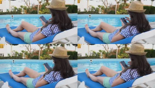年轻漂亮的黑发女子戴着帽子躺在游泳池旁的躺椅上使用手机。3840 x2160 FullHD高清在线视频素材下载