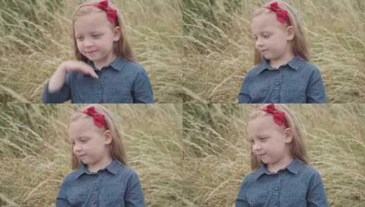 可爱迷人的小女孩，金色头发上系着红丝带，站在秋天的田野里望着远方。漂亮的高加索孩子的肖像享受闲暇。童年和自然的概念。高清在线视频素材下载
