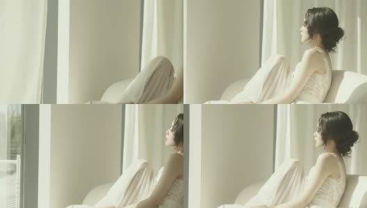 身穿白色婚纱的美丽亚洲新娘坐在落地窗前的沙发上慢镜头拍摄高清在线视频素材下载