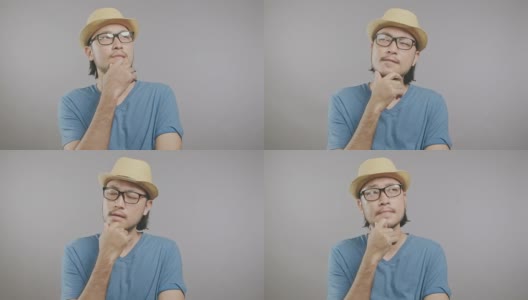年轻的潮人思维孤立在灰色的背景。一名戴着帽子和眼镜的亚洲男子慢慢地将右手放在镜头前的下巴上。中景镜头。高清在线视频素材下载