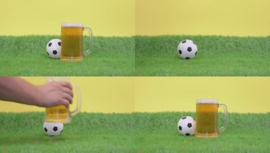一杯清凉的泡沫啤酒站在草坪上，黄色的背景。小玩具足球滚了出来。人拿起杯子解渴，又放回去高清在线视频素材下载