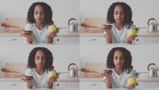 健康的饮食习惯。非裔美国女孩比较甜甜甜圈和熟苹果，选择有机零食的肖像高清在线视频素材下载
